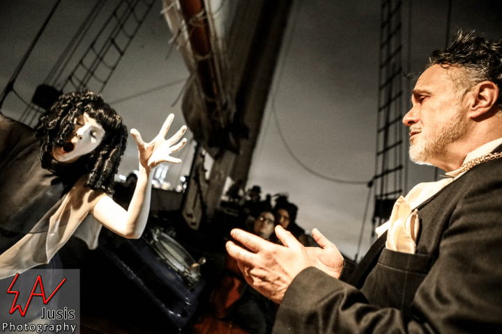 Performer with mime makeup gestures dramatically at a man in a suit on a ship's deck.