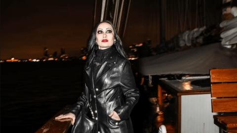 Person in long black coat on a boat at night, city lights in background.