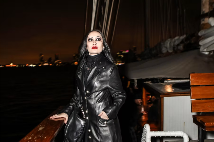 Person in long black coat on a boat at night, city lights in background.