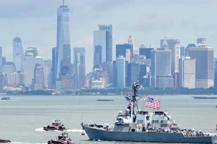 Navy ship with US flag sails past tugboats, New York City skyline in the background.