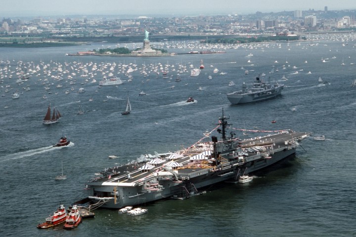 Aerial view of aircraft carrier and boats near a city harbor with the Statue of Liberty in the background.